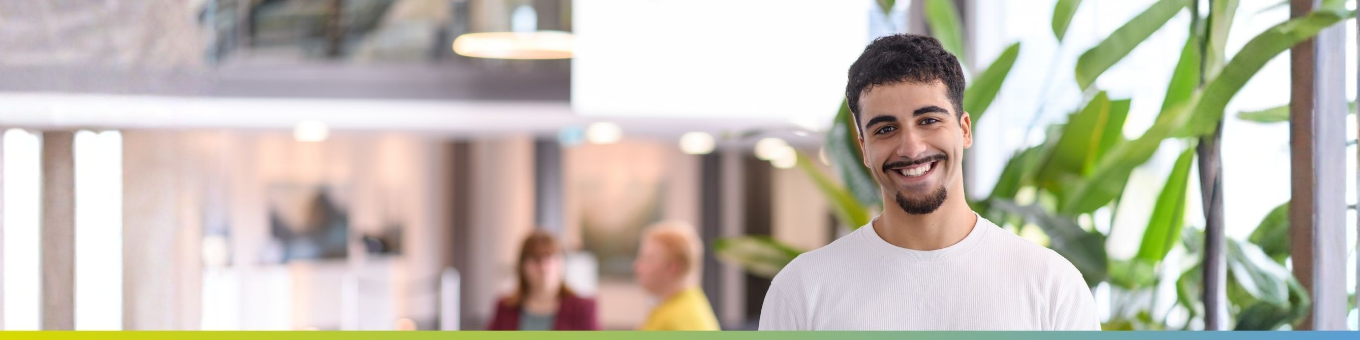 Ein Werkstudent im IT Lab lächelt in die Kamera, zwei Kolleginnen sitzen im Hintergrund