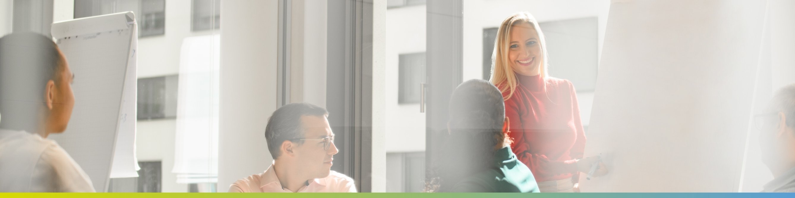 Eine Kollegin steht vor einer Pinnwand mit einem Stift und arbeitet mit mehrere Kolleginnen und Kollegen