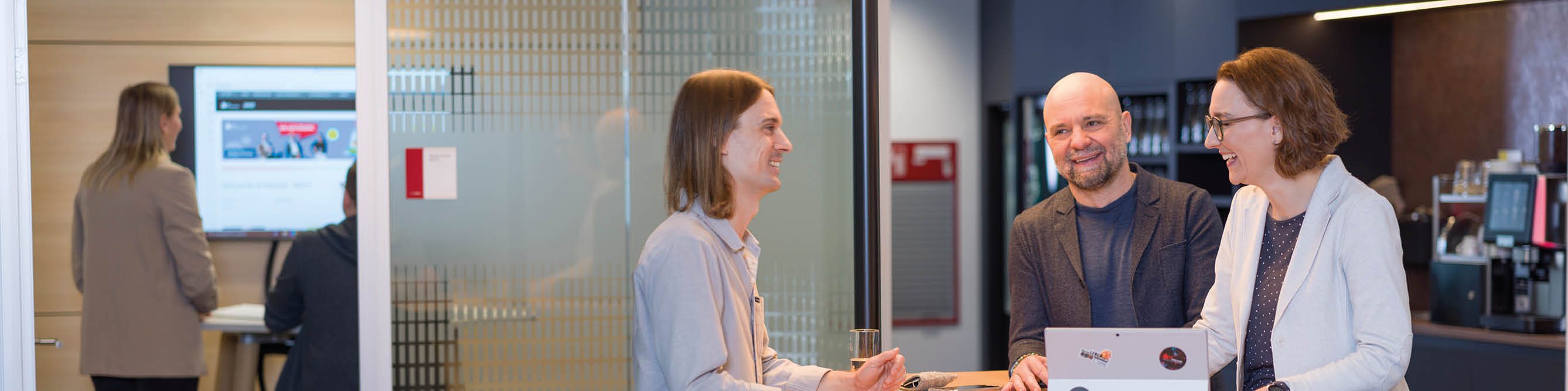 Im Vordergrund arbeitet eine Team stehend an einem Surface, während im Hintergrund zwei Kolleg:innen eine remote Besprechung haben