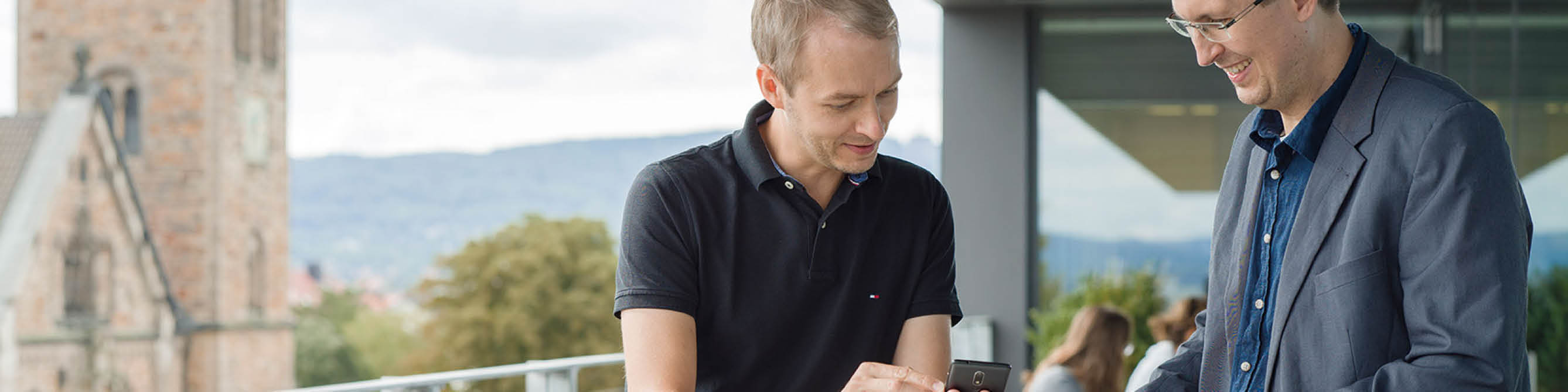 Zwei Kollegen stehen auf der Dachterrasse und schauen gemeinsam auf ein mobiles Endgerät